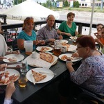 Grupa osób siedzi przy stole na którym stoją talerze z pizzą.