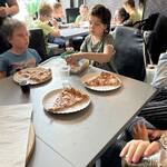 Uczniowie siedzą przy stolikach w pizzerii. Przed nimi stoją talerze z kawałkami pizzy.