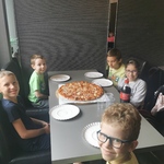 uczniowie siedzą wokół stołu na środku którego stoi duża pizza i talerze.