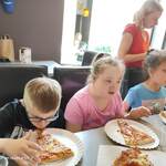 Uczniowie siedzą przy stole na którym stoją talerze z pizzą. Z tyłu stoi nauczycielka.