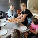 Panie stawiają na stole dużą pizzę. Wokół stołu siedzą dzieci.