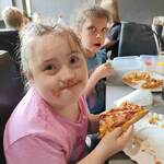 Uczniowie siedzą przy stole i jedzą pizzę. Dziewczynka ma brudną buzię.