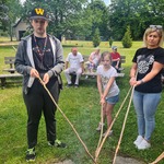 Dwójka uczniów i pani stoją obok ogniska i pieką kiełbaski na długich patykach.