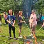 Trójka uczniów stoi przy ognisku i piecze kiełbaski na długich patykach.