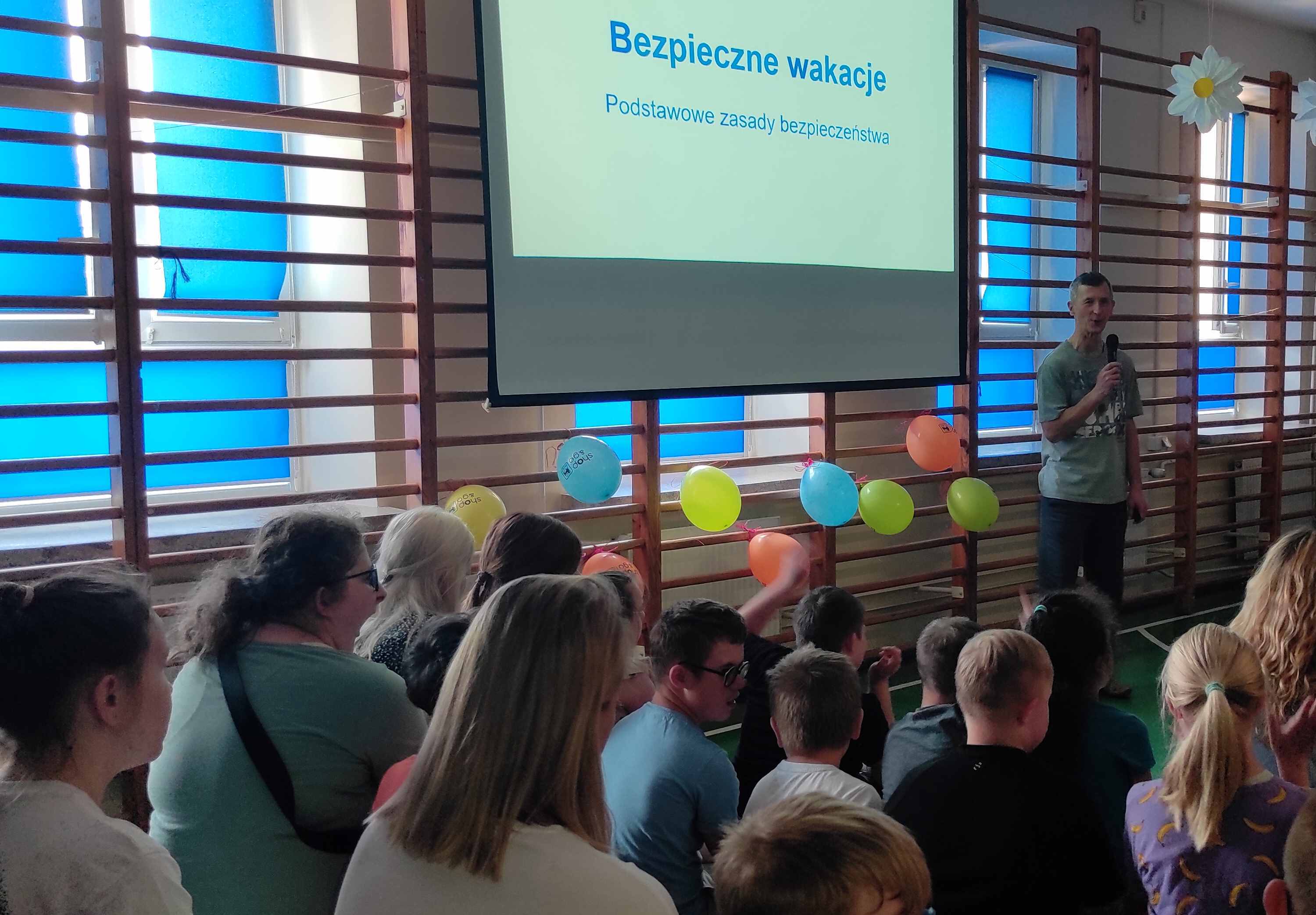 Nauczyciel mówi do mikrofonu. Po bokach siedzą uczniowie,