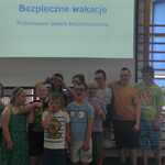 Grupa uczniów na tle tablicy ze slajdem z napisem Bezpieczne wakacje.