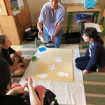 Nauczycielka i uczennice siedzą na dywanie - wykonują dużą pracę plastyczną.