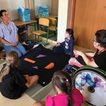 Nauczycielka i uczennice siedzą na dywanie wokół rozłożonej czarnej płachty na której leżą kolorowe elementy.