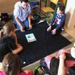 Nauczycielka i uczennice siedzą na dywanie wokół rozłożonej czarnej płachty na której leży jasna kartka.