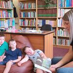 Nauczycielka siedzi na fotelu i czyta książkę. Uczniowie siedzą na pufie. W tle regały z książkami i biurko nauczyciela.