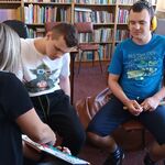 Nauczycielka pokazuje książkę uczniom siedzącym na krzesłach. W tle regały z książkami i stoliki z komputerami.