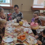Nauczycielka podaje tort uczniom siedzącym wokół urodzinowego stołu.