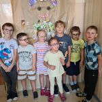 Na zdjęciu stoją uczniowie klasy pierwszych, chłopcy i dziewczynki. Za nimi wisi wykonana przez nich praca. 
Na szarym papierze jest narysowany kontur misia, który jest wypełniony rożnymi materiałami; kartonami, plastikami i folią.