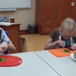 Uczeń i uczennica siedzą za stołem i kroją nożem szczypiorek na desce.
