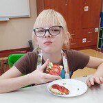 Dziewczynka siedzi przy stole i je kanapkę. Przed nią stoi talerz z drugą kanapką.