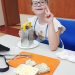 Uczennica siedzi przy stole. Przed nią na talerzu stoi talerz z tarką , leży deska do krojenia z pajdami  chleba oraz stoi pudełko z masłem.