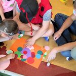 Grupa uczniów siedzi na podłodze i wykonuje prace plastyczną z kolorowych kropek z papieru.