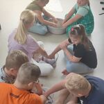 Grupa uczniów siedzi na podłodze i miesza rękoma różowy piasek w miskach.