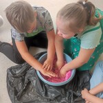 Dwoje uczniów siedzi na podłodze i miesza rękoma różowy piasek w misce
