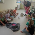 Grupa uczniów siedzi na podłodze. Przed nimi stoją miski z piaskiem. Nauczycielka pochyla się nad wiaderkiem z piaskiem.