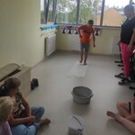 Grupa uczniów siedzi na podłodze. Przed nimi stoją miski z piaskiem.Jeden uczeń idzie po kawałku tkaniny leżącym na podłodze.