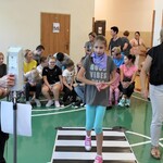 Uczennica przechodzi przez przejście dla pieszych. Obok stoi policjantka. Z tyły siedzą na ławeczkach uczniowie.