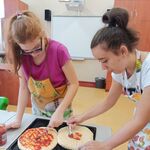 Uczennice smarują keczupem spody pizzy leżące na blaszce.