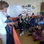 Nauczycielka prezentuje śpiwór. Uczniowie siedzą na krzesłach i na podłodze