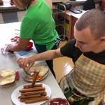 Uczeń siedzi przy stole. Przednim stoją talerze z parówkami i pieczywem. Uczeń trzyma parówkę na widelcu. Obok stoi opiekunka.