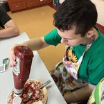 Uczeń siedzi przy stole i nalewa ketchup z butelki na potrawę znajdującą się na talerzu stojącym przed nim na stole.