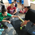 Uczniowie obserwują kolegę wkładającego warzywo do garnka trzymanego przez nauczycielką siedzącą na dywanie.