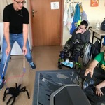 Uczniowie obserwują nauczycielkę poruszającą czarnym pająkiem przywiązanym do kijków,