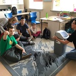Uczniowie siedzący na dywanie obserwują nauczycielkę pokazującą im kosz na śmieci.