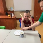 Jeden uczeń siedzi przy stole. Przed nim stoi talerz i kubek.  Drugi uczeń stoi przy stole i dotyka patykiem talerz stojący na stole.