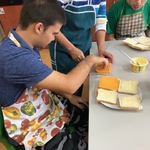 Uczeń siedzi przy stole. Przed nim stoi talerz z tostami. Uczeń bierze plasterek sera z opakowania trzymanego przez opiekunkę.