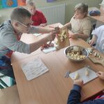 Uczniowie siedzą wokół stołu. Jeden uczeń przekłada pokrojone kawałki owoców do miski stojącej na środku stołu.