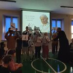 Na tle ekranu ze zdjęciem Jana Pawła II stoi grupa uczniów i ksiądz. Po bokach na ławkach siedzą uczniowie.