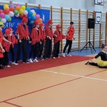 Grupa uczniów w czerwonych ubraniach tańczy na sali gimnastycznej, dziewczynka robi szpagat