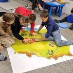 Grupa uczniów siedzi na mapie Polski rozłożonej na podłodze.