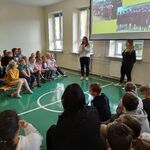 Panie wolontariuszki stoją na tle slajdu wyświetlanego na ekranie. Po bokach siedzą uczniowie naszej szkoły.
