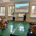 Wolontariuszki stoją na tle slajdu wyświetlanego na ekranie. Po bokach siedzą uczniowie naszej szkoły.