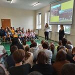 Panie wolontariuszki stoją na tle slajdu wyświetlanego na ekranie. Po bokach siedzą uczniowie naszej szkoły.