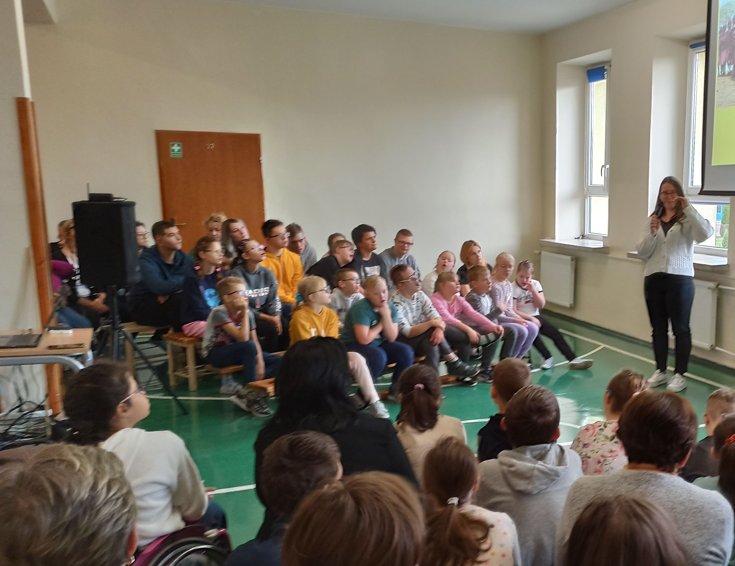 Panie wolontariuszki stoją na tle slajdu wyświetlanego na ekranie. Po bokach siedzą uczniowie naszej szkoły.