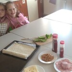 Uczennice siedzą za stołem na którym leżą produkty do wykonania pizzy.