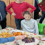Grupa uczniów przygląda się nauczycielce zapalającej świeczki na urodzinowym torcie.