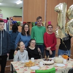 Nauczycielka i grupa uczniów pozują do wspólnego zdjęcia z jubilatką siedzącą przy stole.