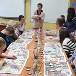 Instruktorka omawia zasady malowania wyrobów z gliny uczniom siedzącym wokół stołu. Przed uczniami leżą na gazetach gliniane figurki.