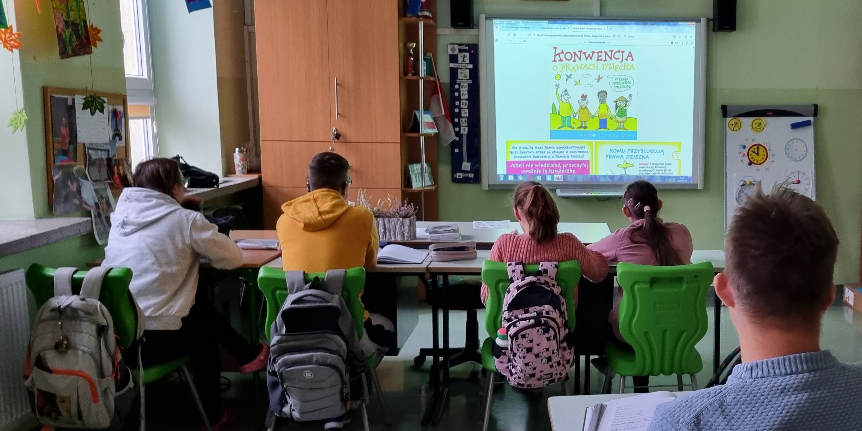 Uczniowie siedzą w ławkach i oglądają prezentację wyświetlana na tablicy interaktywnej.