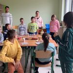Uczniowie stoją wokół stołu i śpiewają solenizantce” 100 lat”.