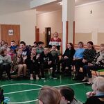 Uczniowie siedzący na ławeczkach słuchają koleżanki śpiewającej do mikrofonu.